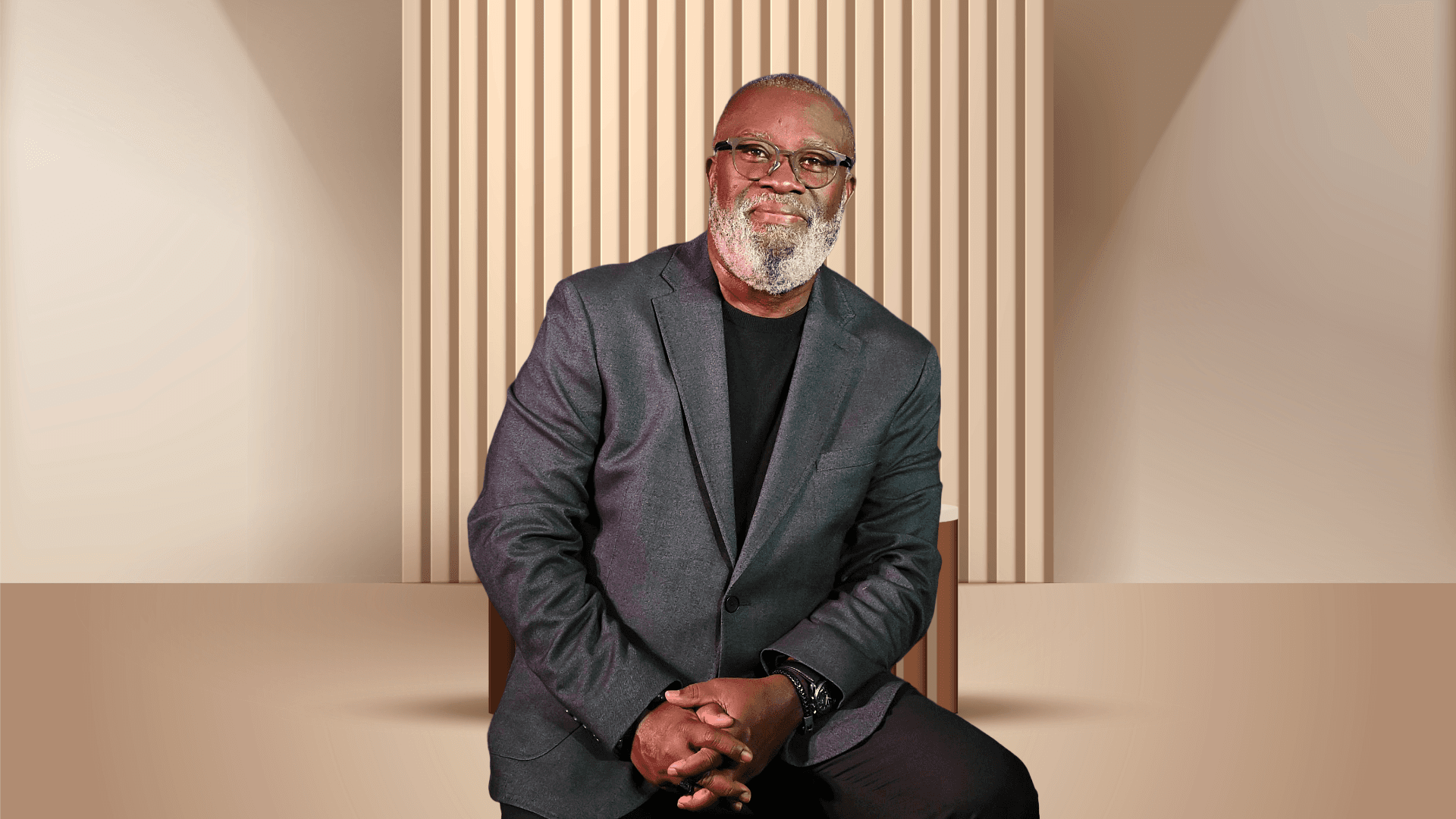 Smiling Black man with grey beard and glasses wearing a grey blazer in a minimalist studio.